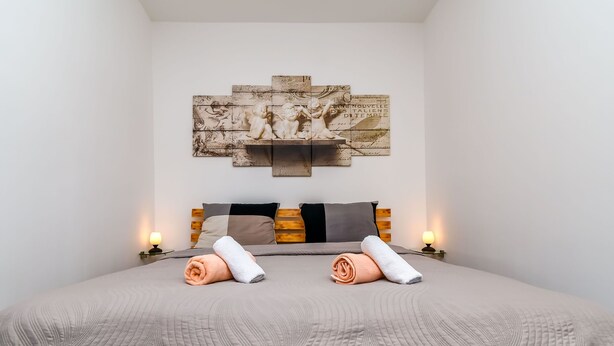 A neatly made bed with a grey quilt, two pillows, and two rolled towels. A wall-mounted artwork above the bed features a relief sculpture. - Apartment Cicko (Trogir)