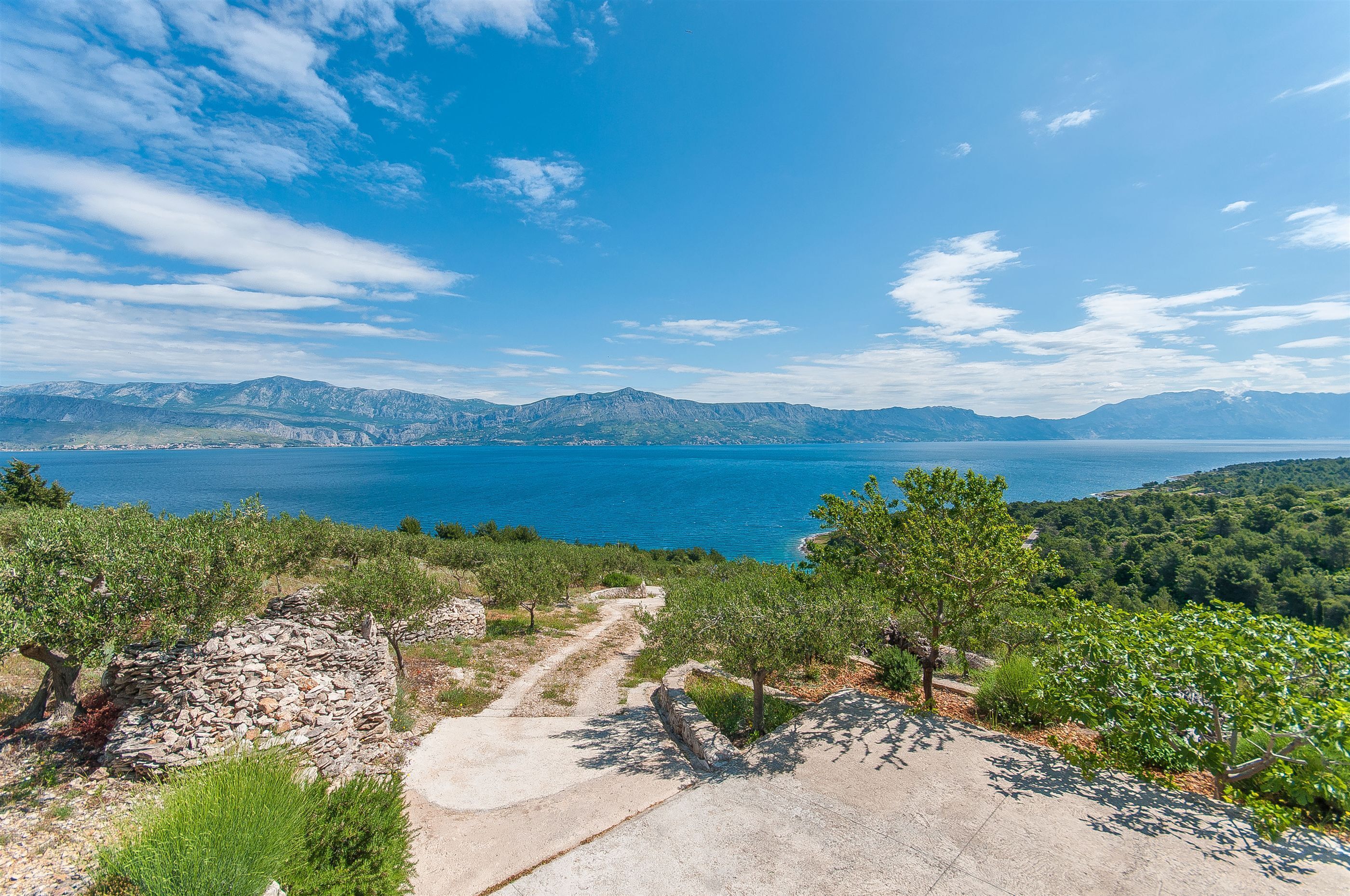 A coastal landscape with a clear sky, a stone pathway, and a view of the sea and mountains.