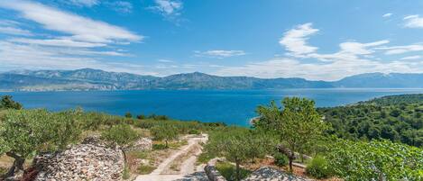 A coastal landscape with a clear sky, a stone pathway, and a view of the sea and mountains.