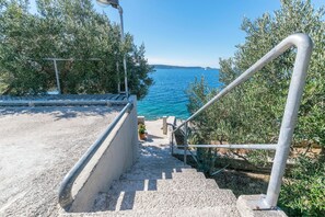 Una escalera de hormigón conduce a una terraza con vistas a un cuerpo de agua, con barandillas de metal y olivos.