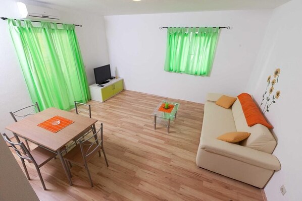 A living room with a wooden floor, a beige sofa with orange cushions, a glass coffee table, a TV on a green cabinet, and green curtains. - Apartments Branka (Podgora)