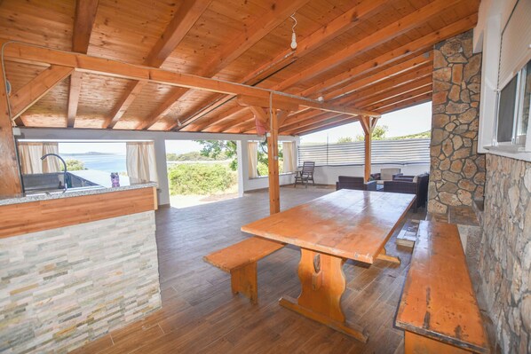 A spacious covered patio with a wooden table and benches, stone walls, and a view of the outdoors. - House Romano (Pasman)