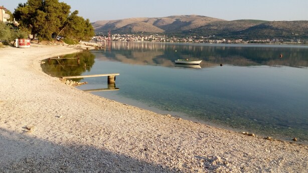 Ein Kiesstrand mit einer Holzbank, ruhigem Wasser, einem kleinen Boot und weit entfernten Hügeln.