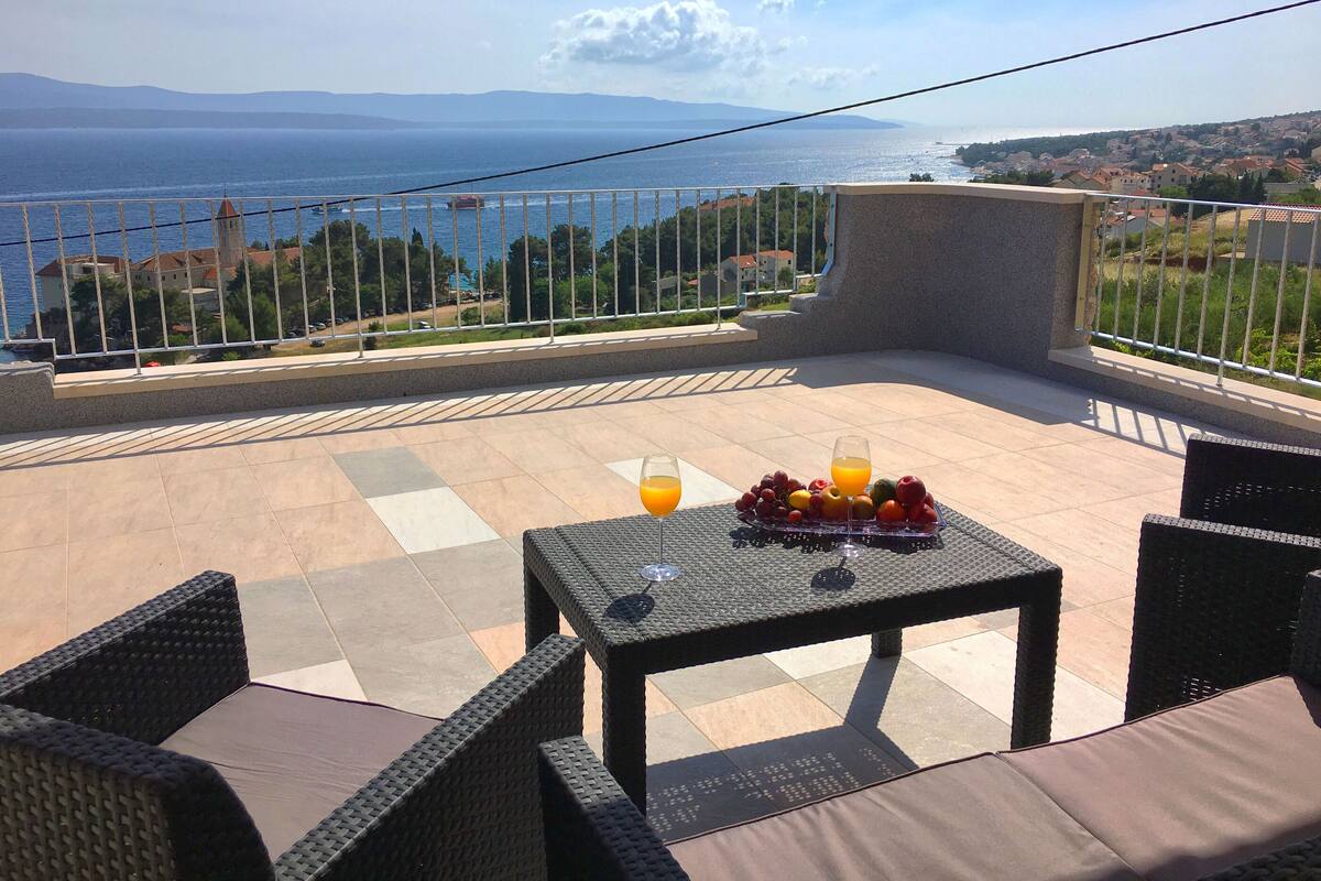 Una terraza con una mesa preparada para dos personas, vista al mar y un pueblo lejano.