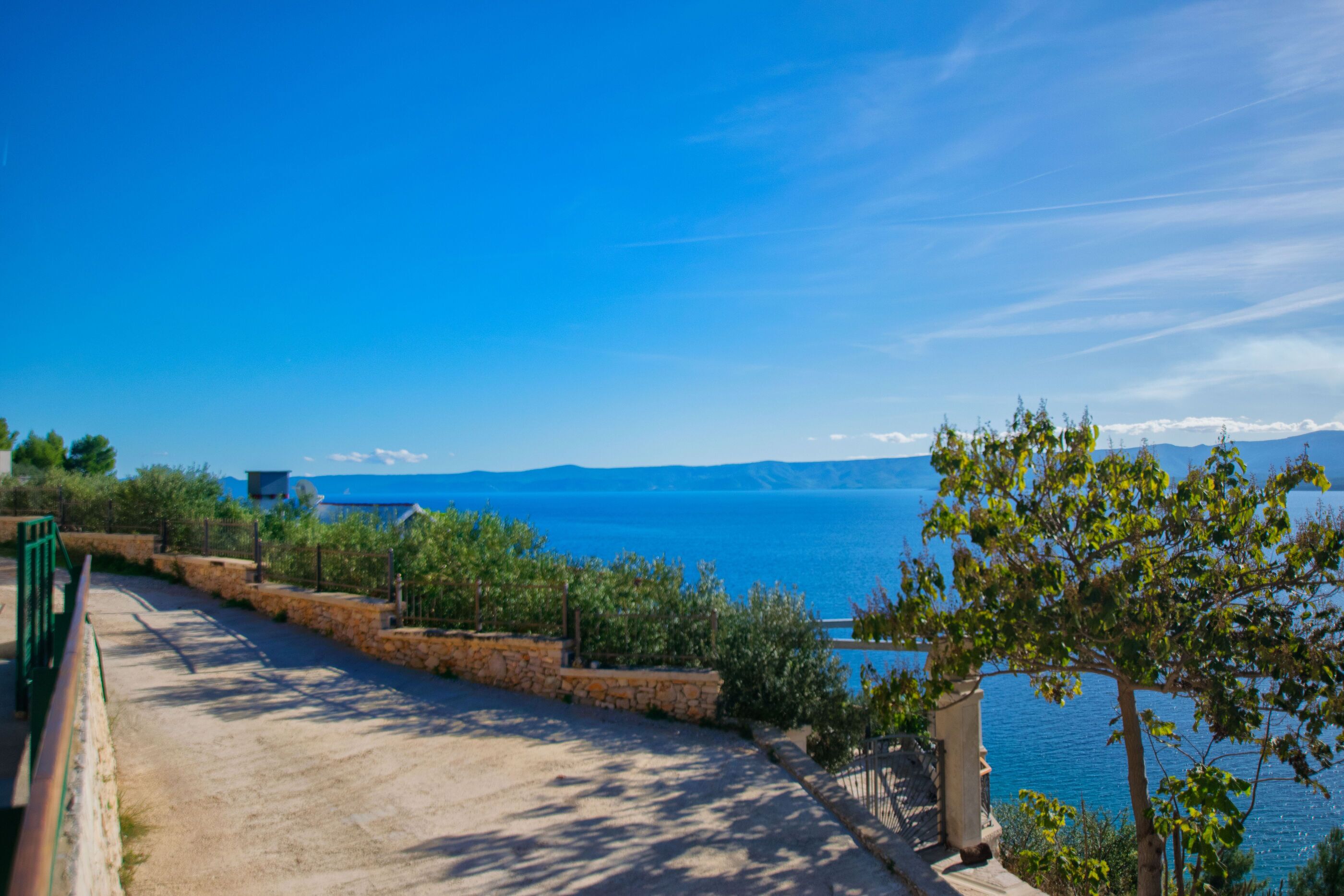 A coastal road with stone walls and greenery, leading to a view of the sea and distant mountains.