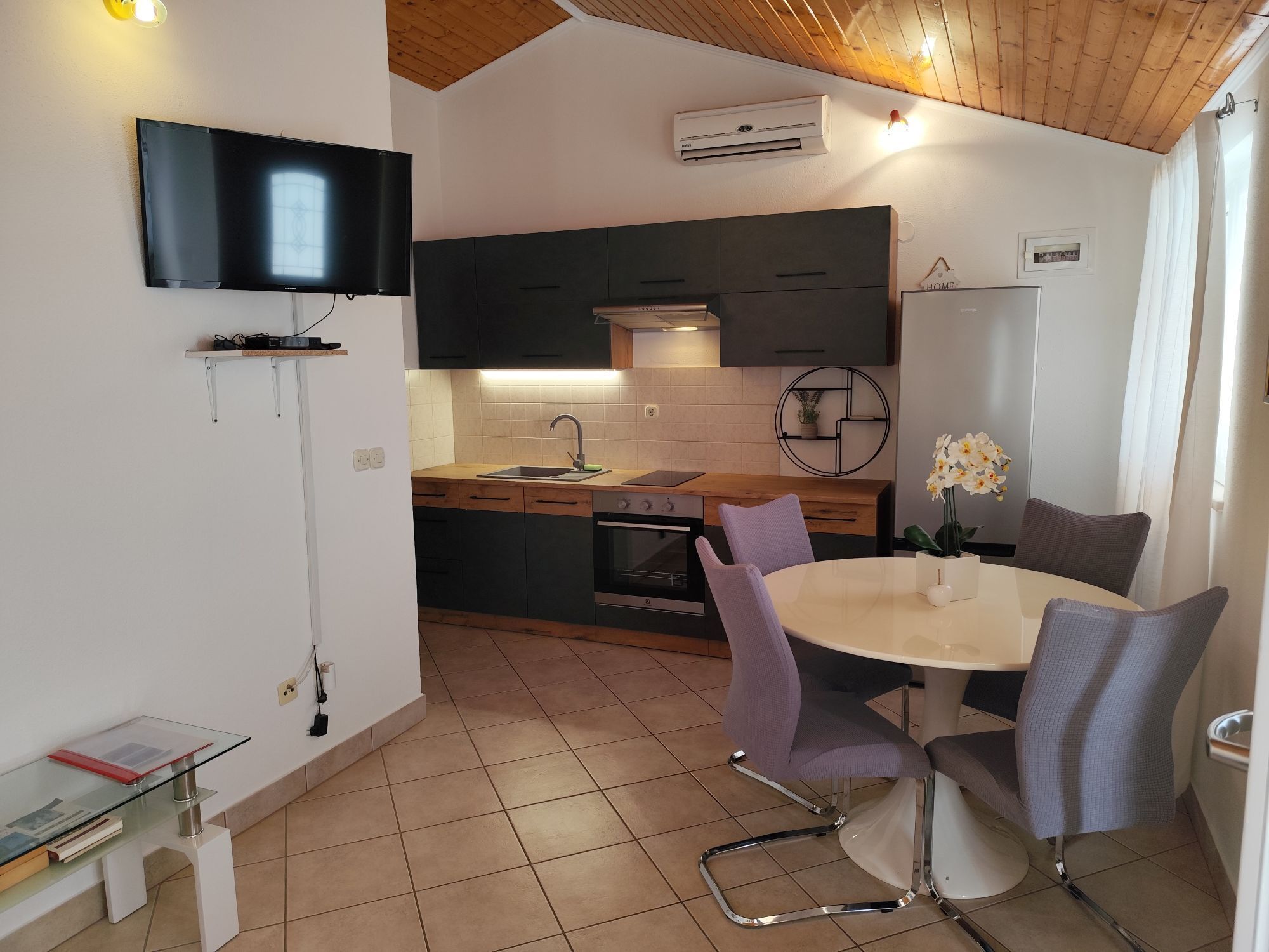 A modern kitchen with a built-in oven, a dining table with chairs, and a TV mounted on the wall.