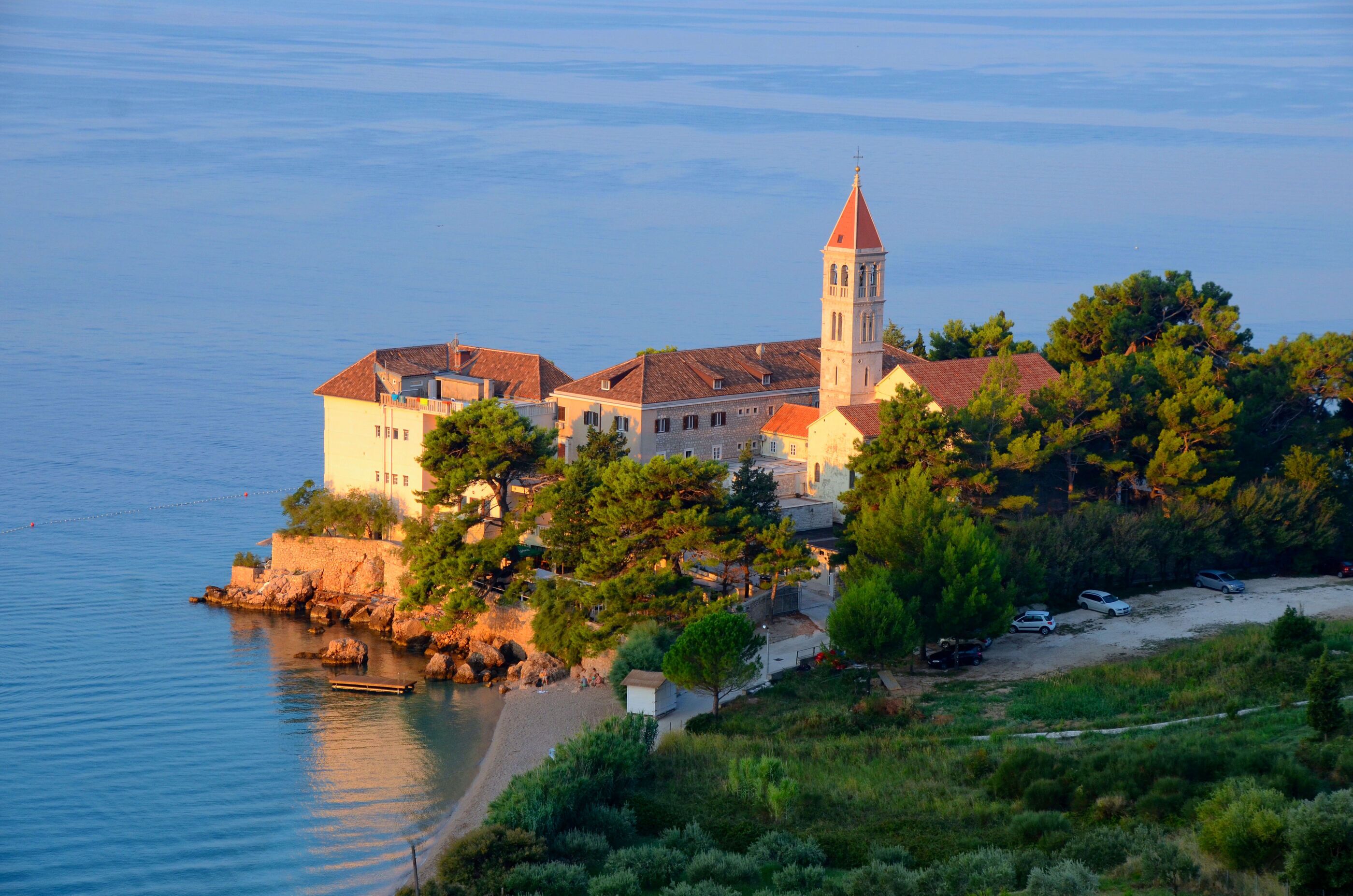 A coastal building with a prominent tower, situated on a rocky shoreline with a beach and parked cars.