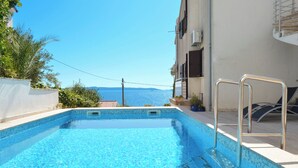A swimming pool with clear blue water, a metal ladder, and a view of the sea in the background. - Apartments Inge (Seget)