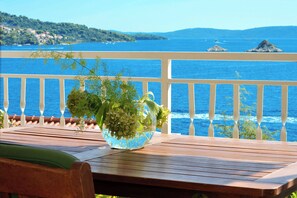 A wooden table with a glass vase containing green plants, overlooking a coastal view with a railing and distant islands. - Apartments Inge (Seget)