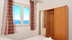 A bedroom with a bed, a window with curtains, a wooden wardrobe, and a view of the sea. - Apartments Inge (Seget)