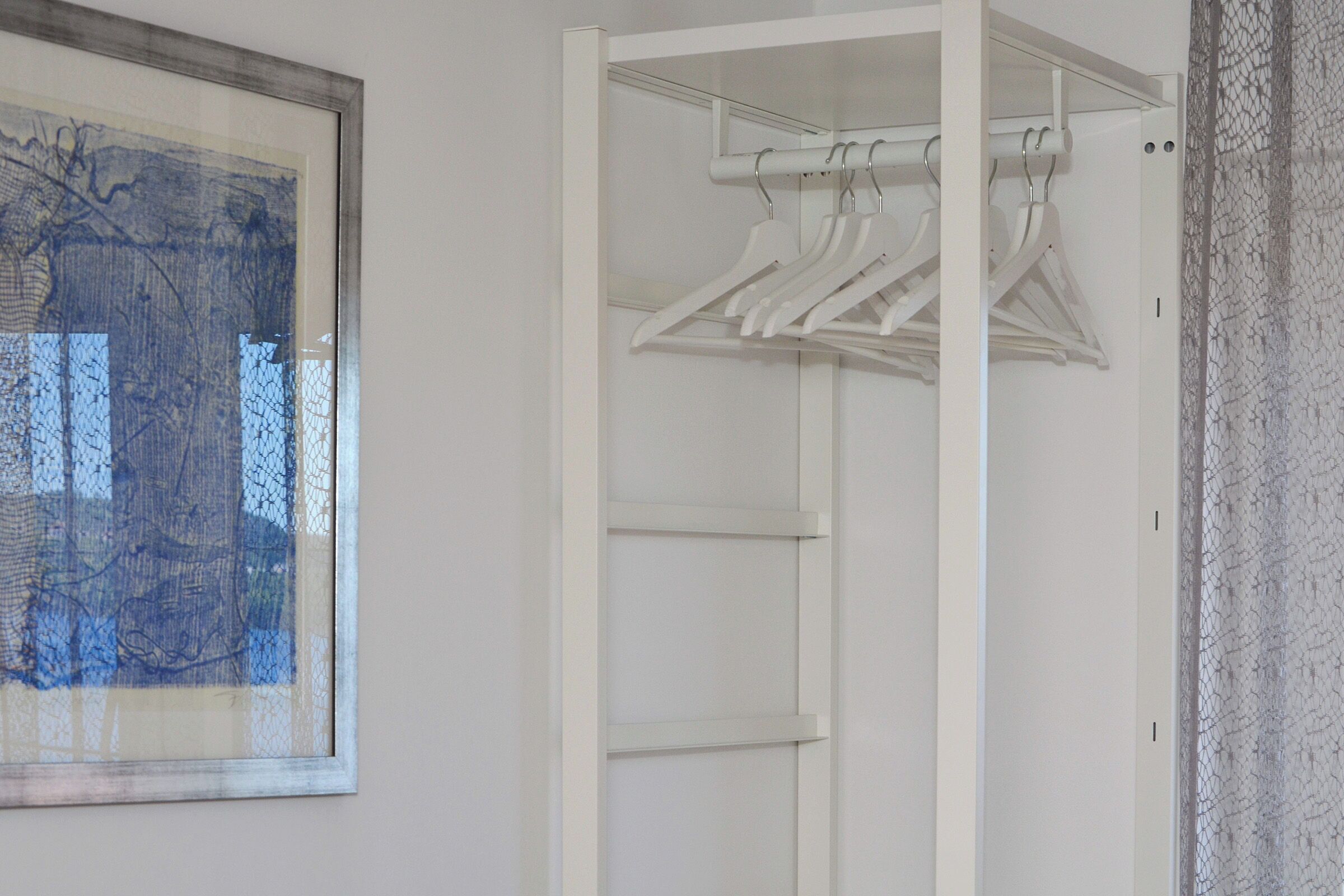 A white closet with multiple shelves and hanging space, a framed abstract painting on the wall, and a textured wall on the right.