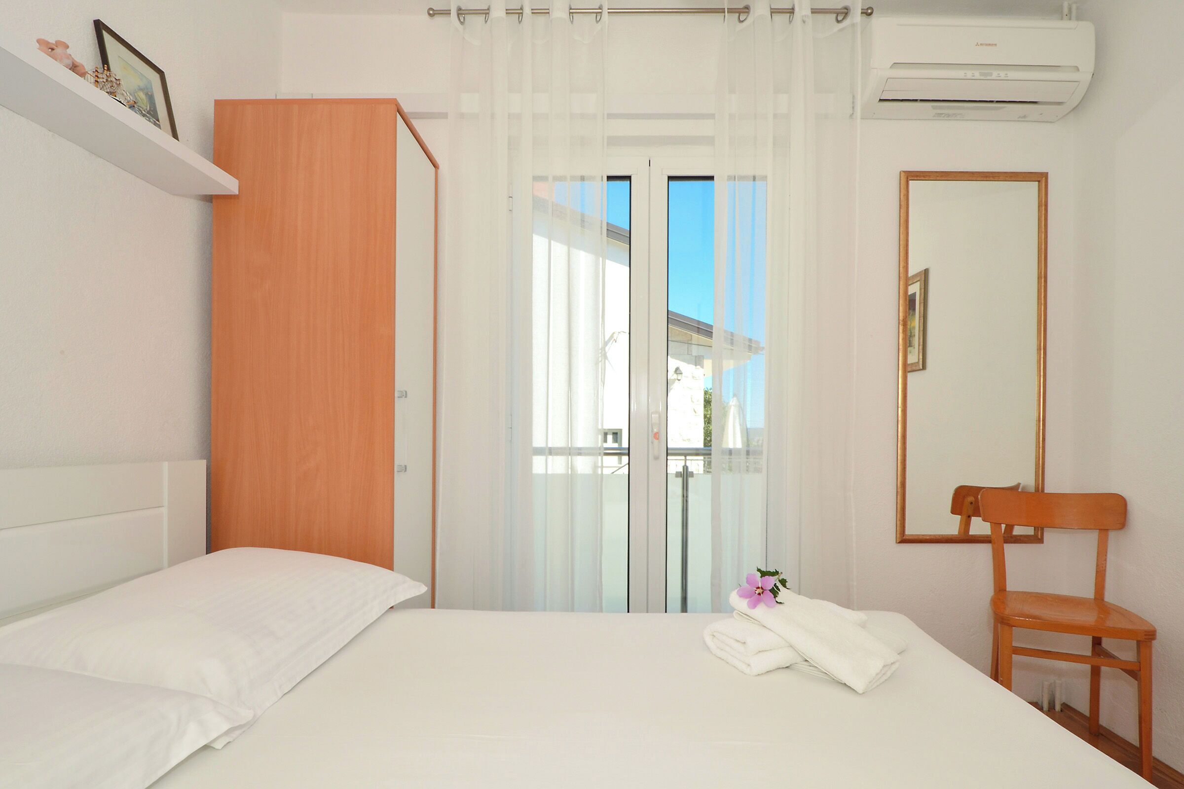 A hotel room with a bed, a wooden wardrobe, a chair, a bathroom with a glass door, and an air conditioning unit.