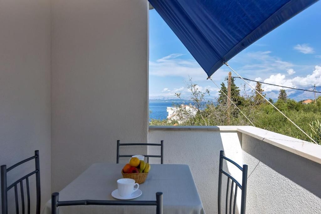 A balcony with a table set for two, a basket of fruit, and a cup. The view includes a body of water and distant buildings.
