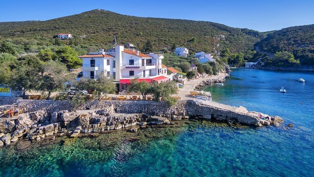 Un bâtiment situé sur la côte, avec plusieurs étages, un restaurant au rez-de-chaussée et une eau d’un bleu limpide en premier plan.