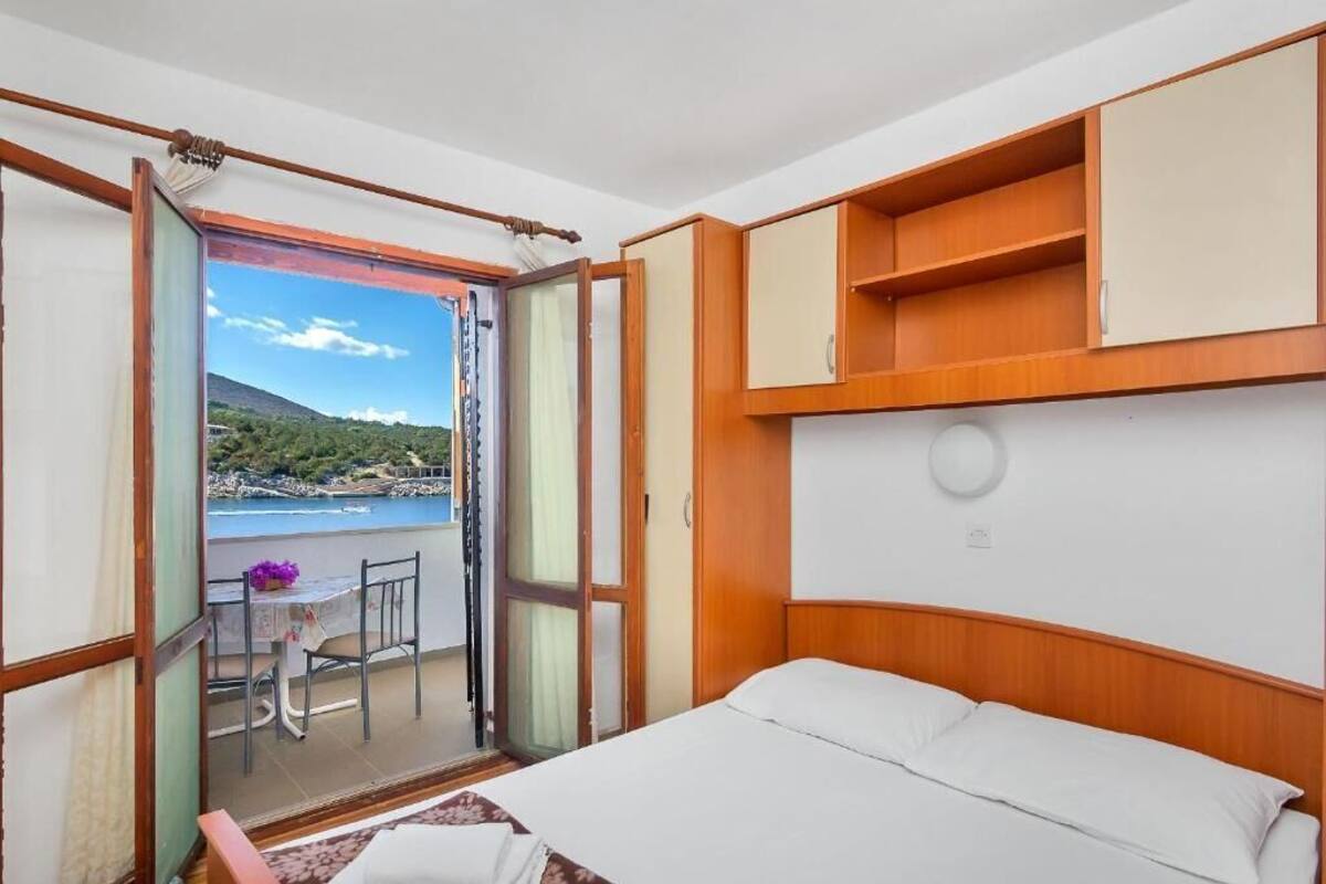 Ett sovrum med en säng, en sänggavel i trä, ett litet bord med en stol och en balkong med utsikt över havet.