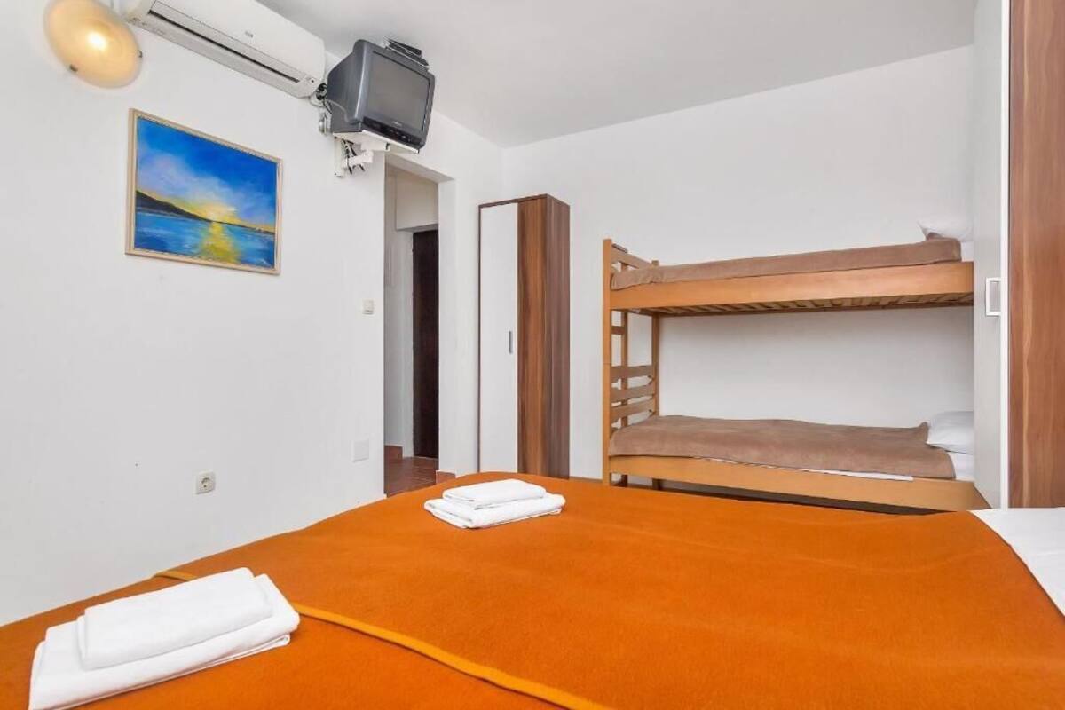 Ett rum med en våningssäng, en vanlig säng med ett orangefärgat överkast, en tv som är monterad på väggen och en tavla som föreställer en solnedgång.