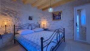 A bedroom with a stone wall, a bed with a patterned bedspread, two bedside lamps, and a painting on the wall. - House Bjankura (Smokvica)