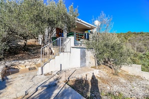 Una casa moderna con una escalera de piedra que conduce a un porche cubierto, rodeada de olivos y un cielo azul despejado.