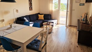 A living room with a sofa, a dining table with chairs, a small round table, and a view of the outdoors through a glass door. - Apartment Petra (Rab)