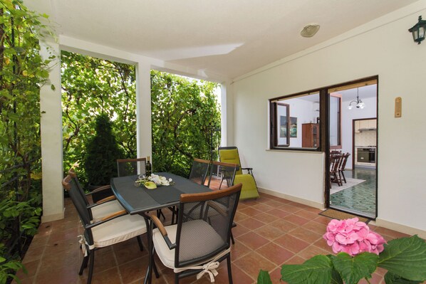 A patio with a table and chairs, surrounded by greenery and a view into a kitchen area. - Apartment Lorenzo (Seget)