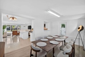 A modern kitchen with white cabinetry, a dining area with a wooden table and chairs, and a large window providing natural light. - Evergreen Retreat Home for 7 (Oakland Park)