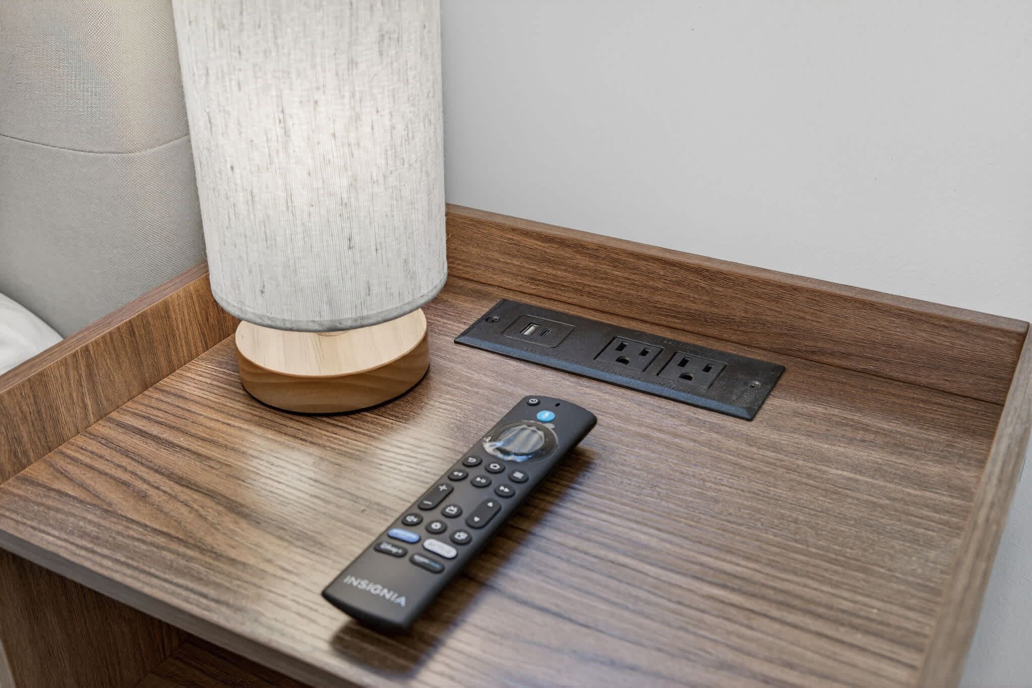 A wooden bedside table with a lamp, a remote control, and a power outlet.