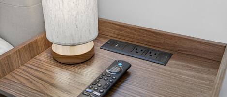 A wooden bedside table with a lamp, a remote control, and a power outlet.