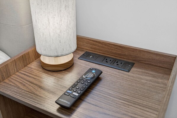 A wooden bedside table with a lamp, a remote control, and a power outlet. - Evergreen Retreat Home for 7 (Oakland Park)