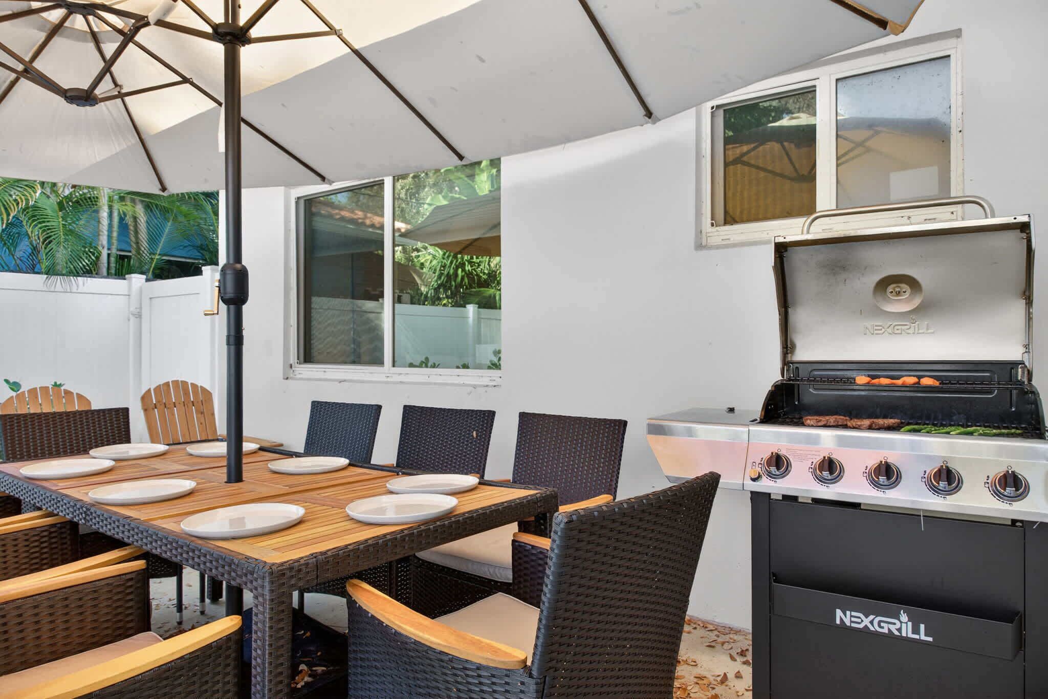 Un patio al aire libre con una mesa de comedor dispuesta para seis personas, rodeada de sillas de mimbre. Se ve una parrilla a gas en el lado derecho.