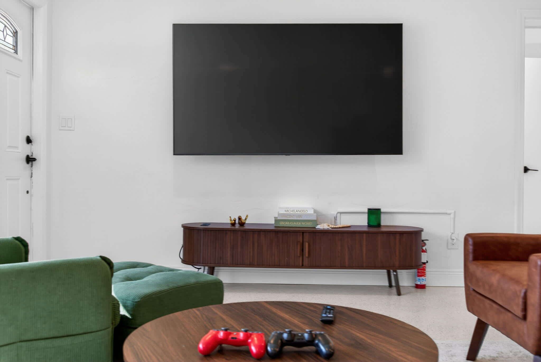 A modern living room with a large flat-screen TV mounted on the wall, a wooden cabinet with decorative items, a green sofa, a brown leather armchair, and a round coffee table with two game controllers.