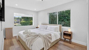 Une chambre dotée d’un grand lit, de deux tables de chevet avec des lampes, d’un téléviseur fixé au mur et de grandes fenêtres donnant sur un paysage verdoyant.