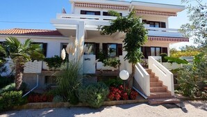 Una moderna casa a due piani con facciata bianca, tetto in tegole rosse e un giardino curato con diverse piante e fiori.
