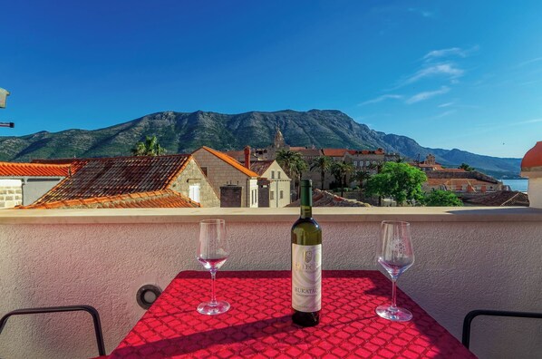 A table with a bottle of wine and two glasses on a red tablecloth, overlooking a mountainous landscape with buildings.