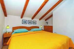 Un dormitorio con cabecera de madera, cama con colcha naranja, dos almohadas turquesa, una mesita de noche con lámpara y un aparador.