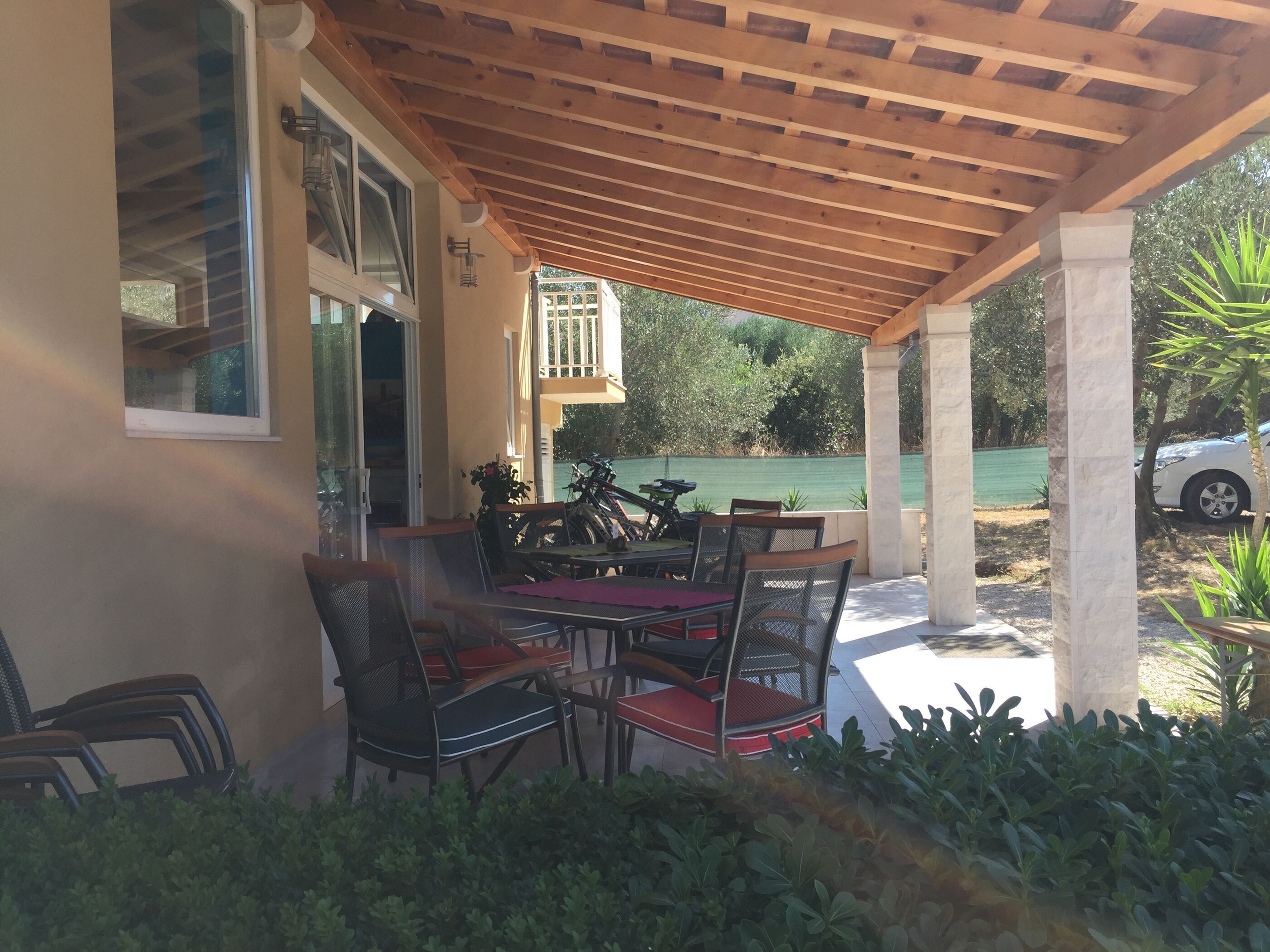 Een terras buiten met een houten pergola, een tafel met stoelen, en uitzicht op een groene hekken en bomen.