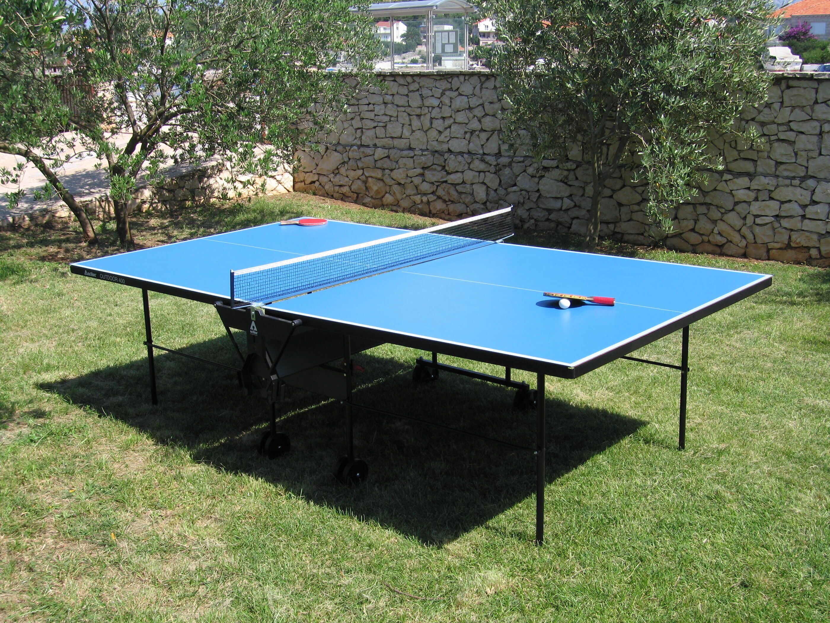 A blue ping pong table with a net, set up on a grassy lawn with a stone wall and trees in the background.