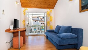 A room with a blue sofa, a wooden desk with a TV, a pink ottoman, and a balcony with yellow curtains. - Apartments Family Corner (Vodice)