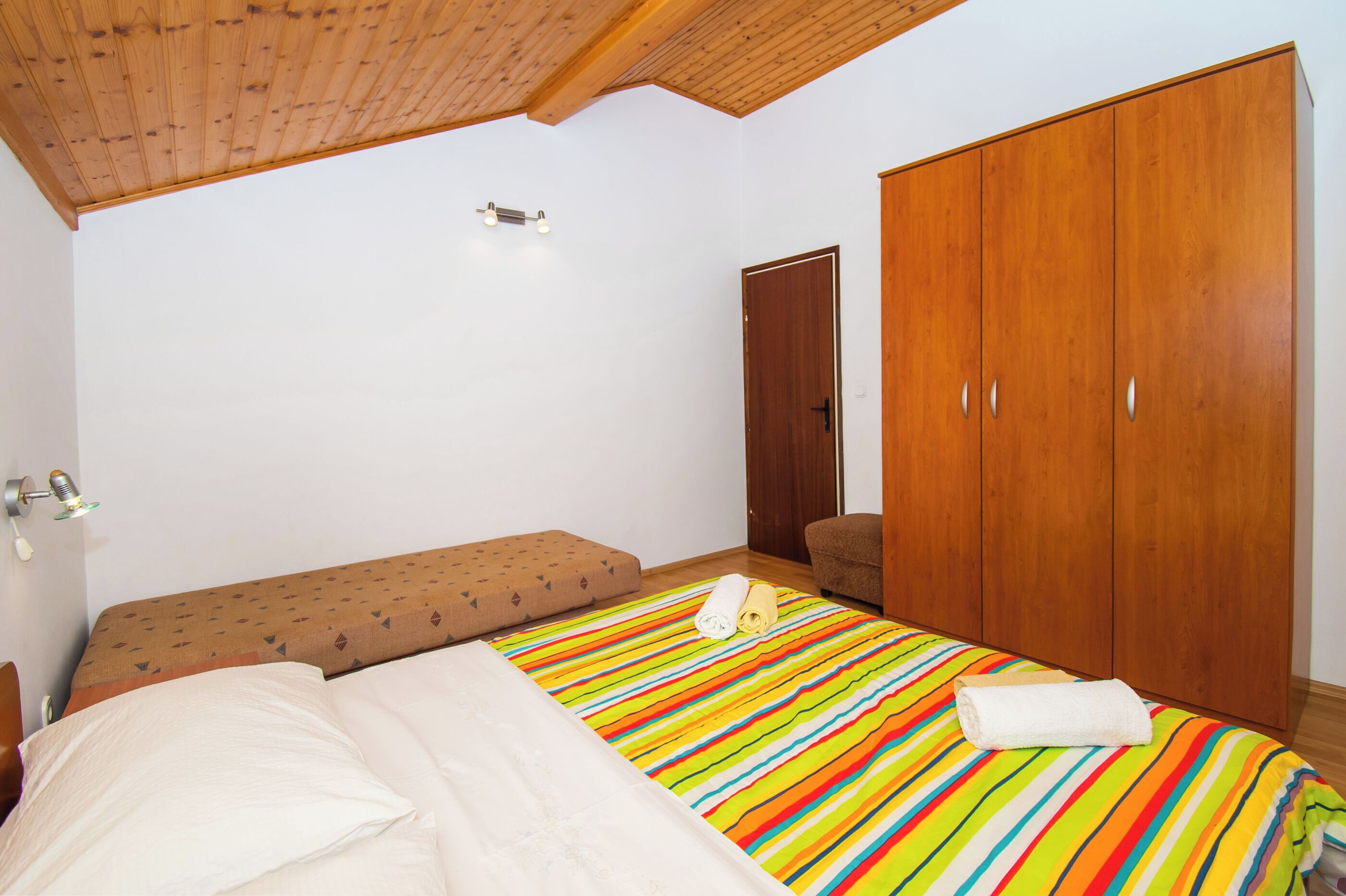 Een slaapkamer met een houten plafond, een bed met een gestreept beddegoed, een bankje, een kledingkast en een deur.