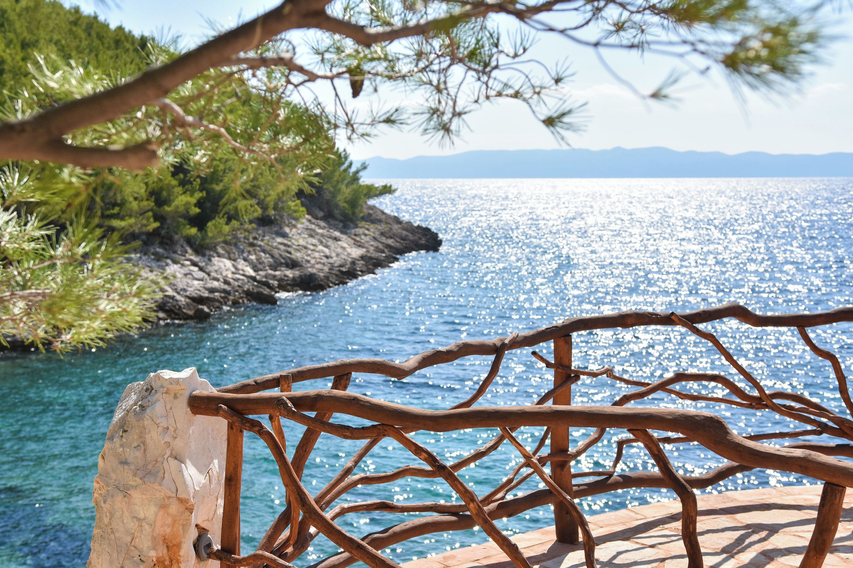 A wooden railing overlooking a clear blue sea with rocky shores and greenery.