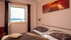Un dormitorio con cama, ventana con cortinas, lámpara de pared y un cuadro de un atardecer.