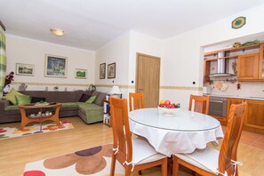 A living room with a sofa, a glass dining table, and wooden chairs. There is a kitchen area with cabinets and a stove. - Apartment MM (Okrug)