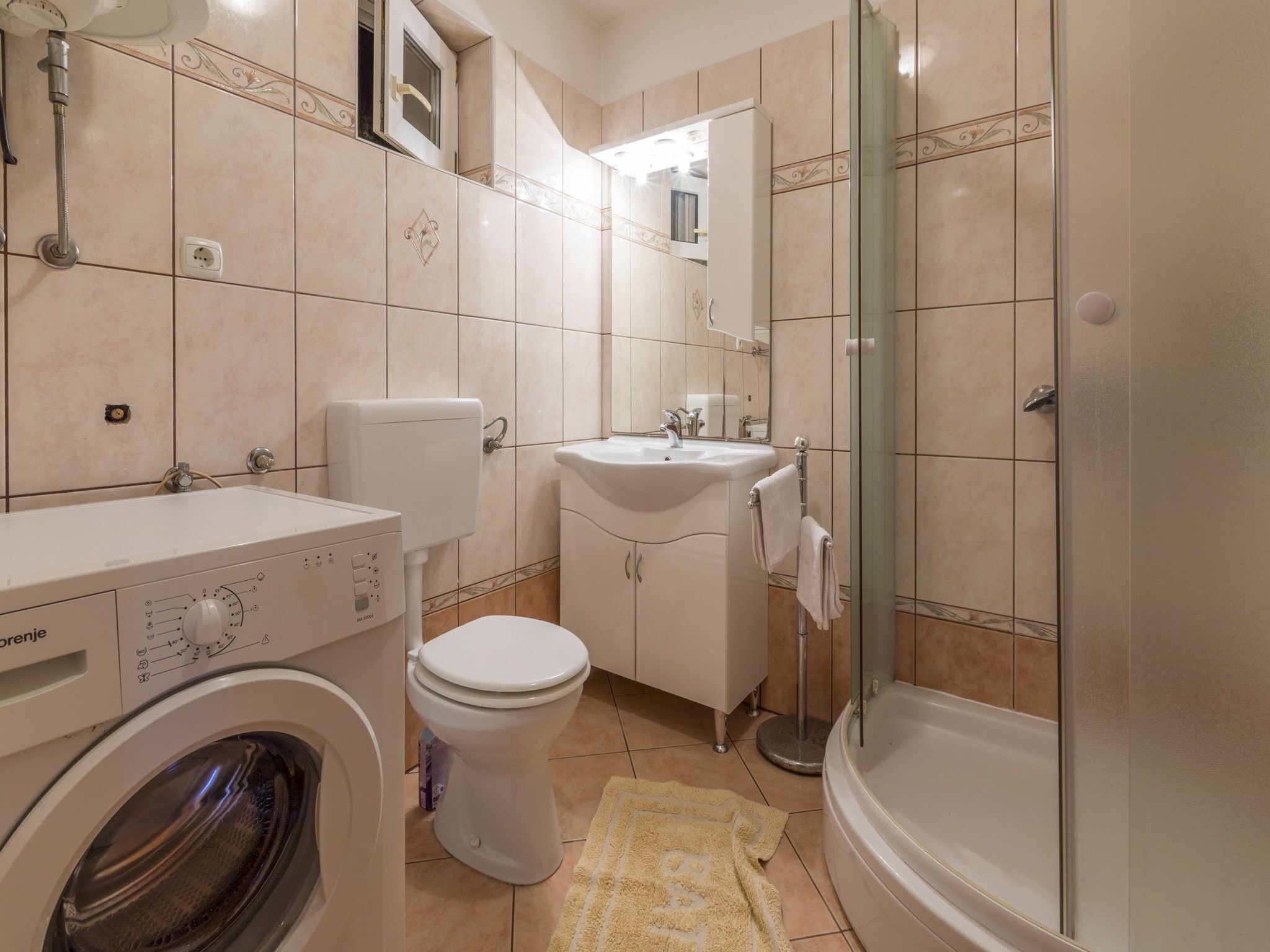 Une petite salle de bain équipée d’un lave-linge, de toilettes, d’un lavabo et d’une douche à parois de verre.