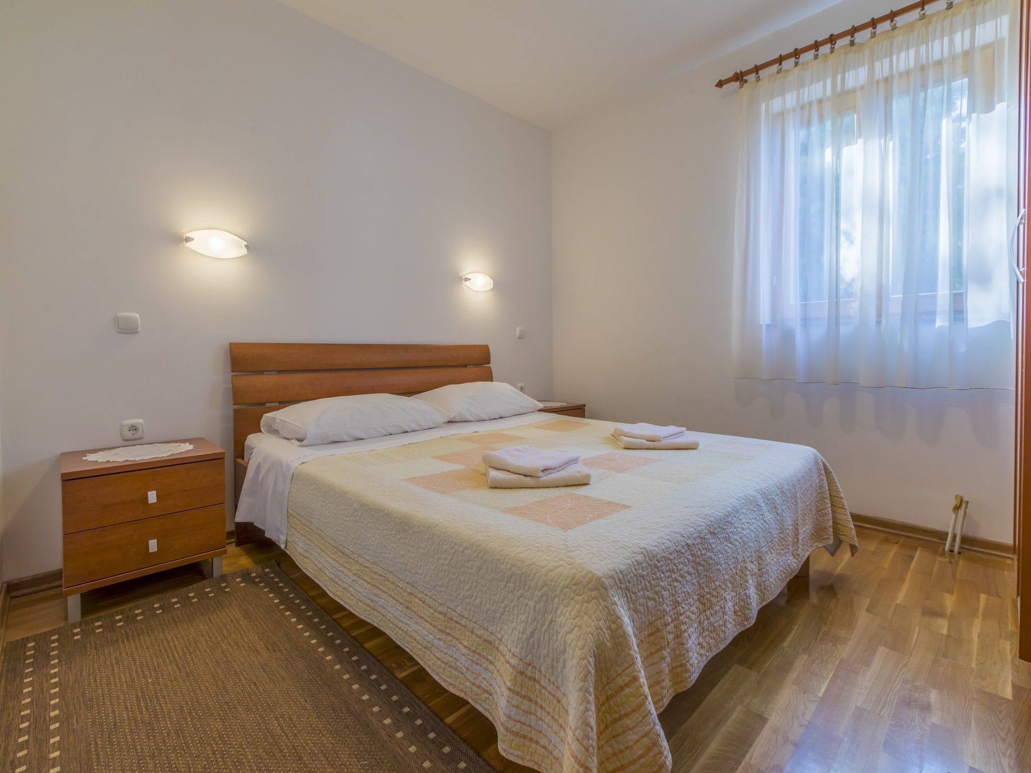 A bedroom with a wooden bed, two bedside tables, a nightstand, a patterned rug, and a window with sheer curtains.