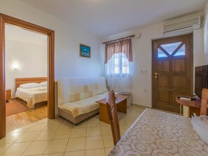 A hotel room with a bed, a sofa, a small table, a desk, a chair, a window with curtains, a door, and an air conditioning unit. - Apartments Dino (Crikvenica)