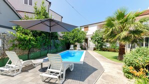 A pool area with lounge chairs and a large umbrella, surrounded by greenery and a house. - Apartment Milena (Vodice)