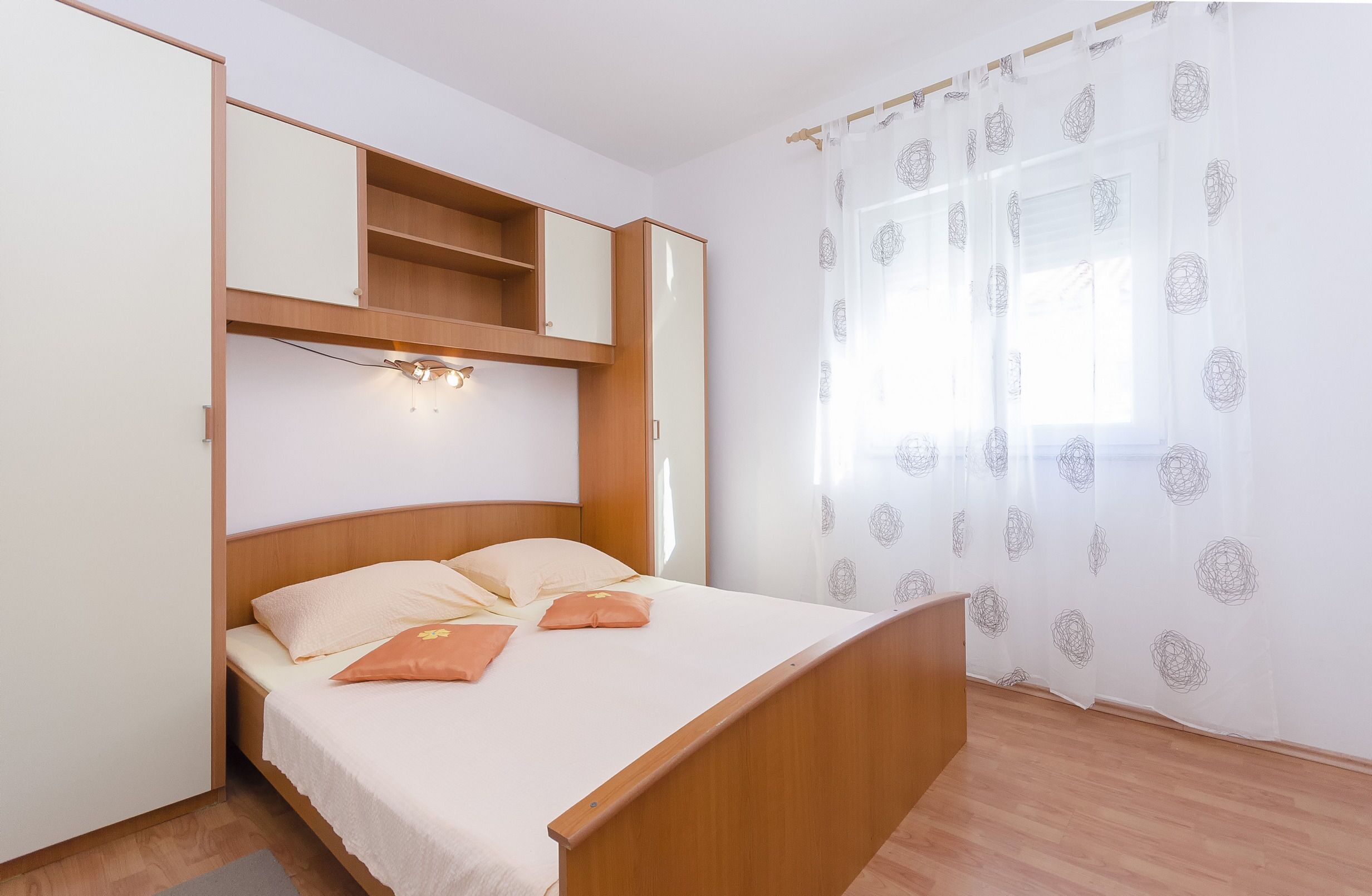 A bedroom with a bed, a wardrobe, a shelf, and a window with patterned curtains.