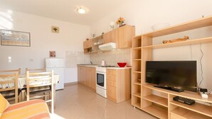 A compact kitchen with a refrigerator, stove, and microwave. There's a dining table with chairs and a TV on a wooden stand. - Apartment Zdravko (Vodice)