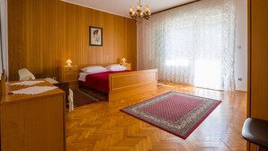 A bedroom with a wooden floor, a bed with white and red bedding, two bedside tables with lamps, a wooden dresser, and a large window with sheer curtains. - Apartment Darinka 1 (Crikvenica)