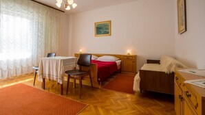 A room with a wooden herringbone floor, a bed with a red blanket, a wooden nightstand with a lamp, a chair, a small table with a white tablecloth, and a wooden dresser. - Apartment Darinka 1 (Crikvenica)