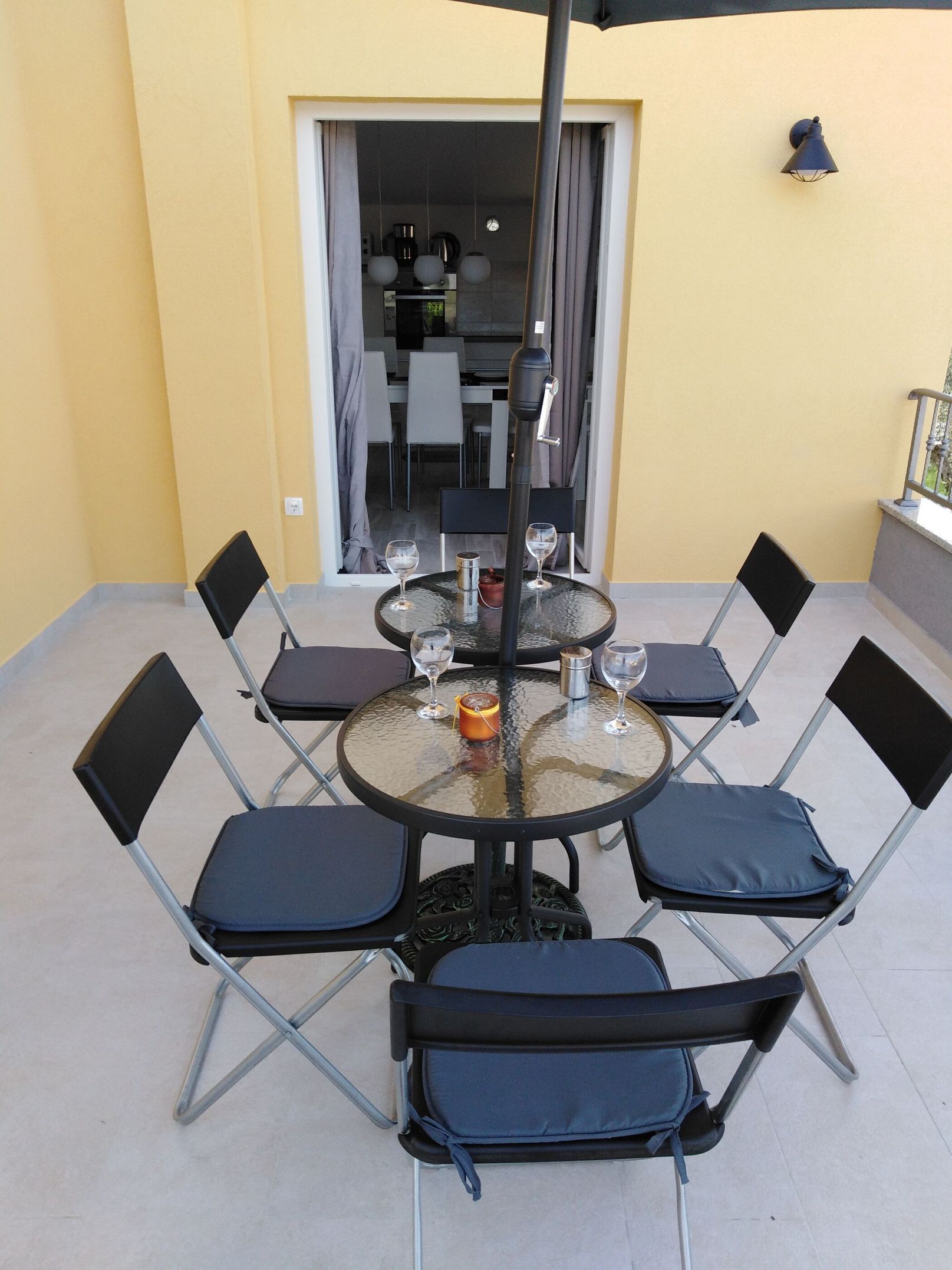 A round glass table with six chairs, a candle, and two wine glasses on a balcony with yellow walls and a view into the dining area.